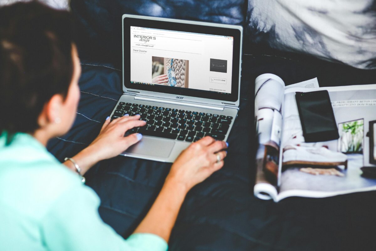 A Woman On A Bed Browsing And Blogging With A Laptop And A Magazine Nearby.