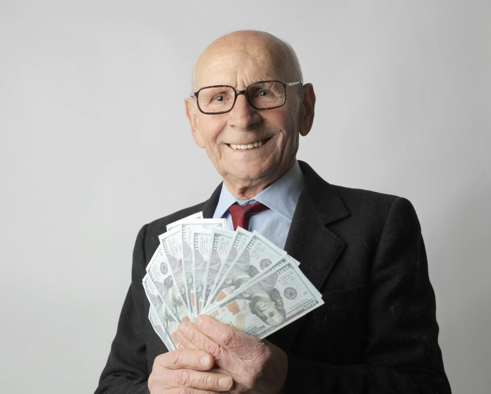 Senior Man In A Suit With Glasses Holding Us Dollars, Symbolizing Wealth And Savings.